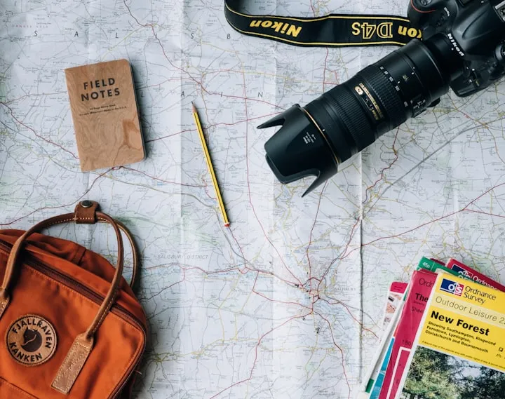Overhead arrangement of a Nikon DSLR, orange Fjallraven backpack, Field Notes notebook, and maps on a paper topographic map