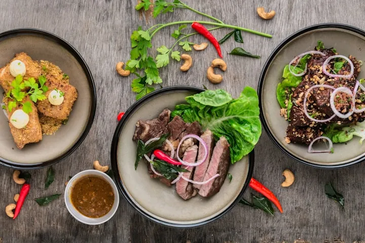 Top-down view of three gourmet bowls with grilled meat, fried cubes, herbs, and chili on a rustic wooden table