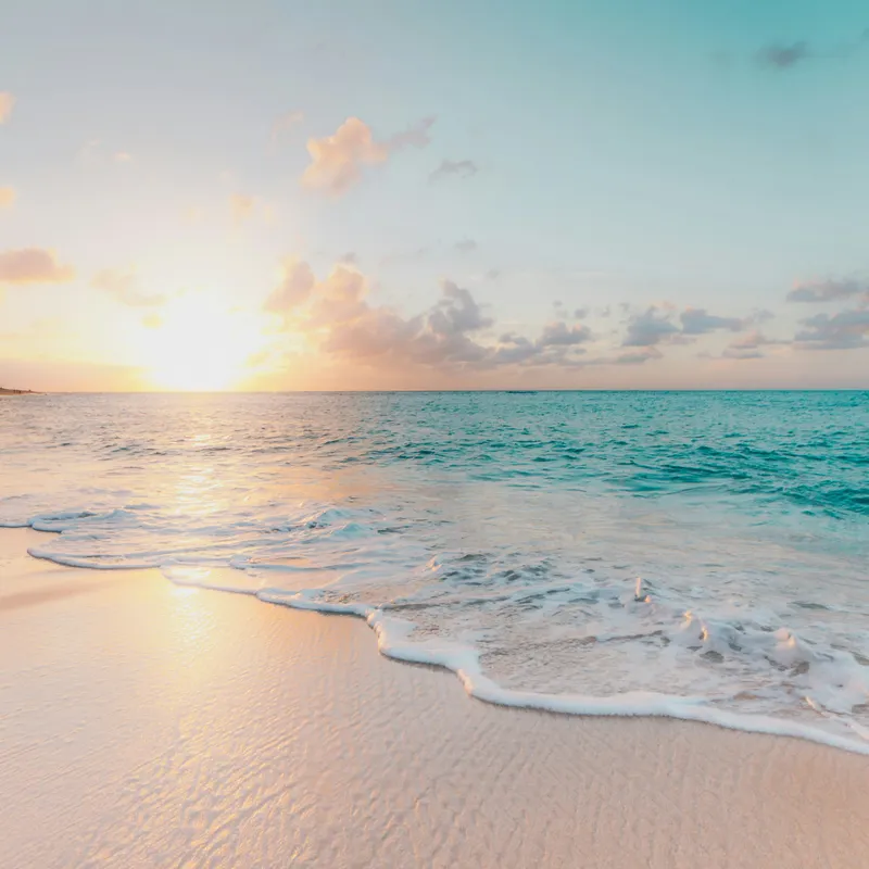 Tropical beach at sunset with a gentle wave on white sand and turquoise water under a soft orange sky