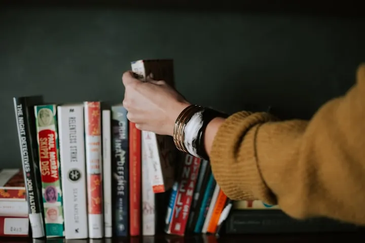 Hand in a mustard-yellow sweater reaching for a book on a dark shelf among crime and fiction titles