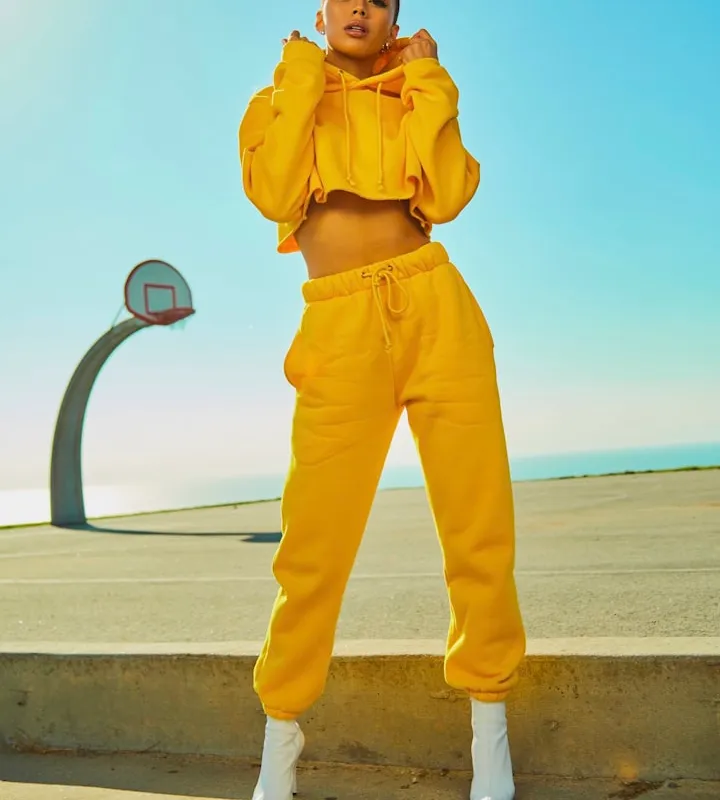 Woman in a bright yellow cropped hoodie and matching joggers posing on an outdoor basketball court by the ocean