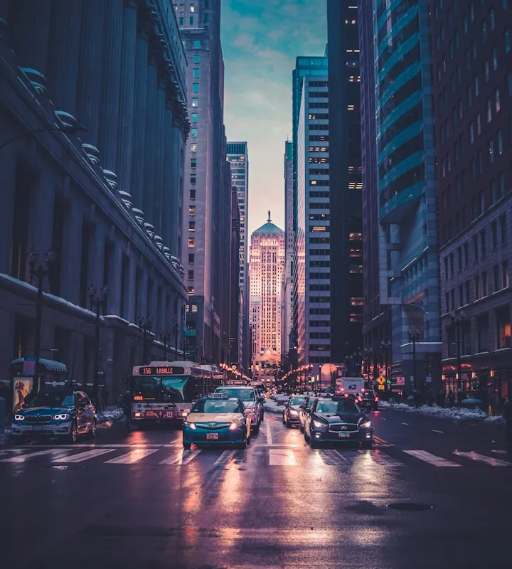 Vertical view down a wet city street at dusk with skyscrapers, a bus, and headlights reflecting on the pavement