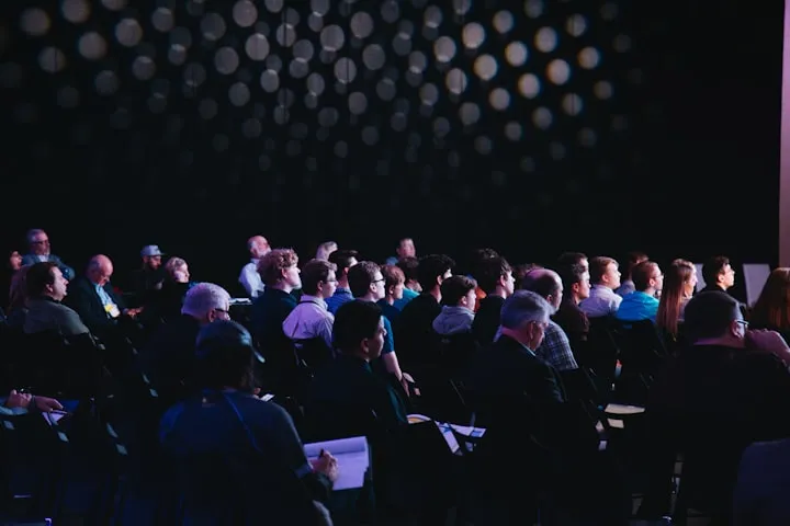 Seated audience in a darkened conference hall looking toward a presentation