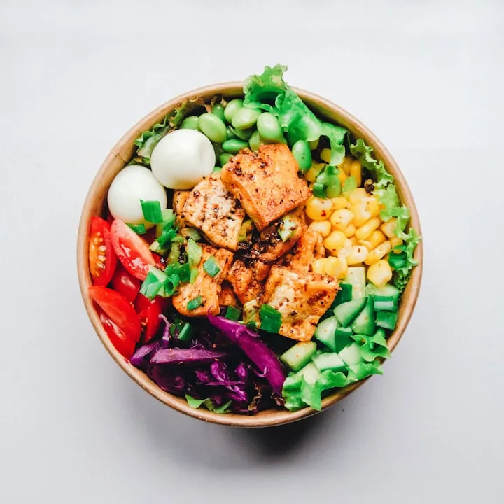 Top-down colorful Buddha bowl with seared tofu, corn, cucumber, red cabbage, tomatoes, and quail eggs