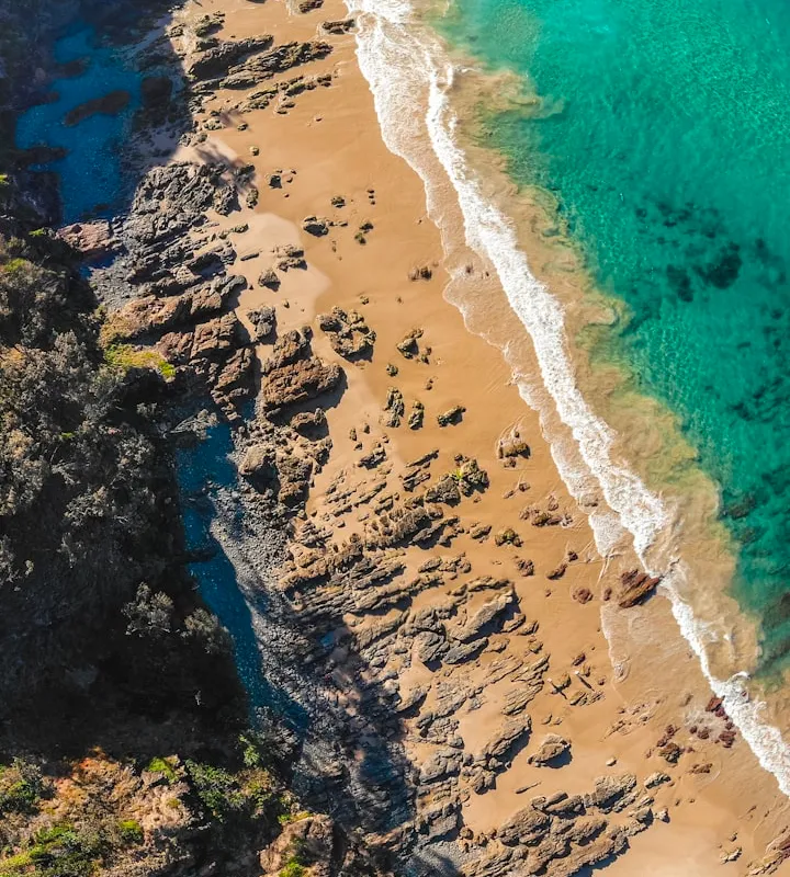 Top-down aerial of turquoise ocean meeting a rocky, sandy shore beside dark cliffs