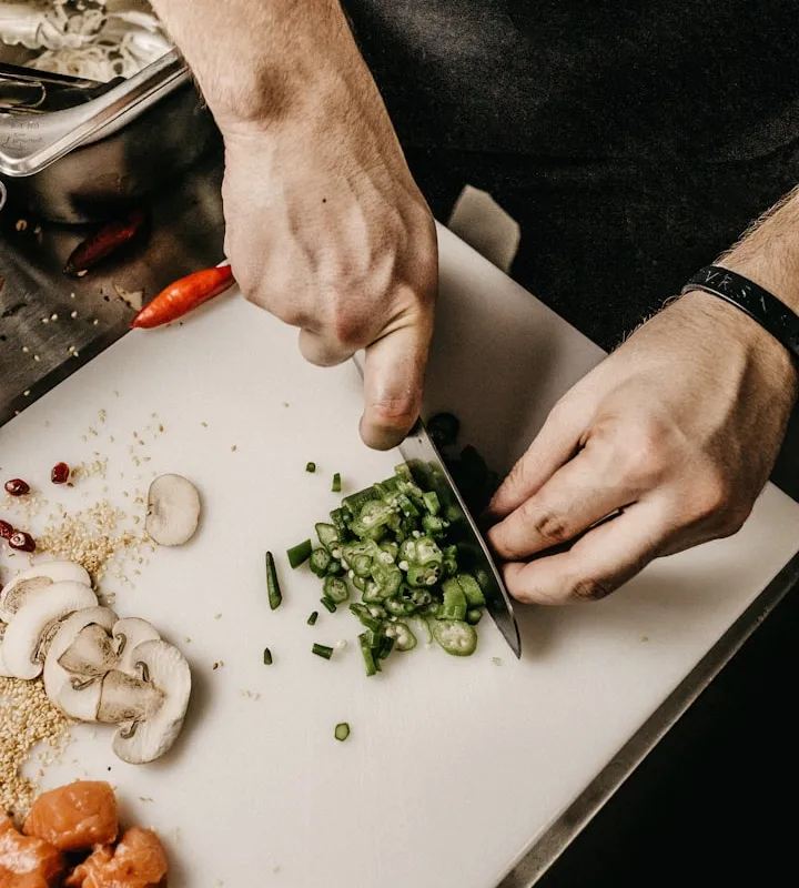 Top-down view of a chef's hands chopping green chili on a white board beside sliced mushrooms and cubed salmon