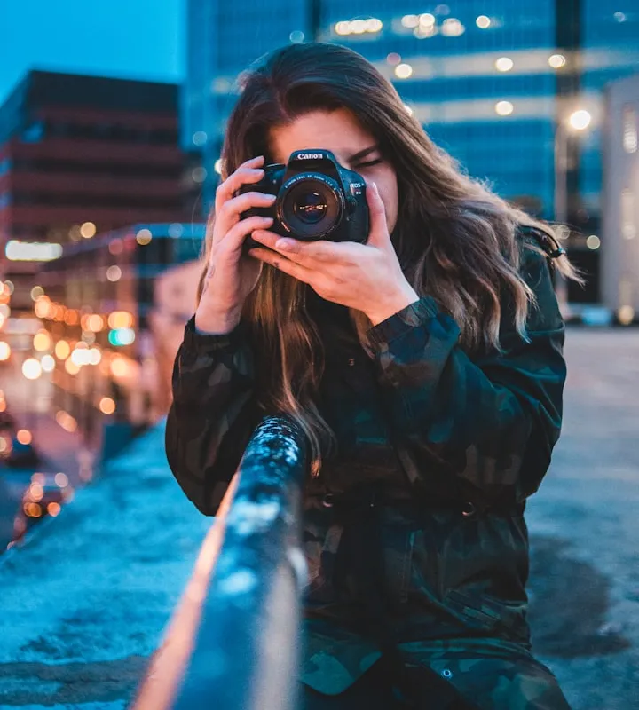 Woman in a camouflage jacket holding a Canon DSLR to her eye against a blurred city night backdrop
