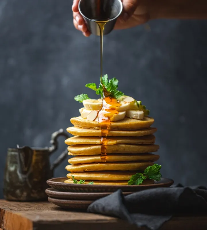 Tall stack of pancakes topped with banana and mint, with amber syrup being poured from a small metal cup