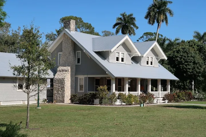 Two-story gray house with white trim, a stone chimney, and palm trees under a bright blue sky